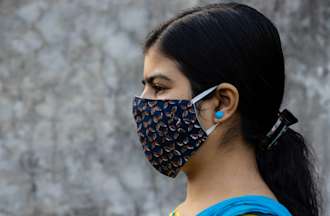 [Featured Image]:  A female contact tracer, wearing a blue vest, a yellow patterned top and a face covering.  She is standing in front of a building with a white wall.  