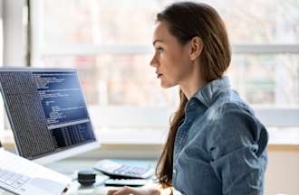 Une personne en chemise bleue est assise à un bureau près d'une fenêtre et travaille sur un code à l'aide d'un ordinateur portable et d'un second écran.