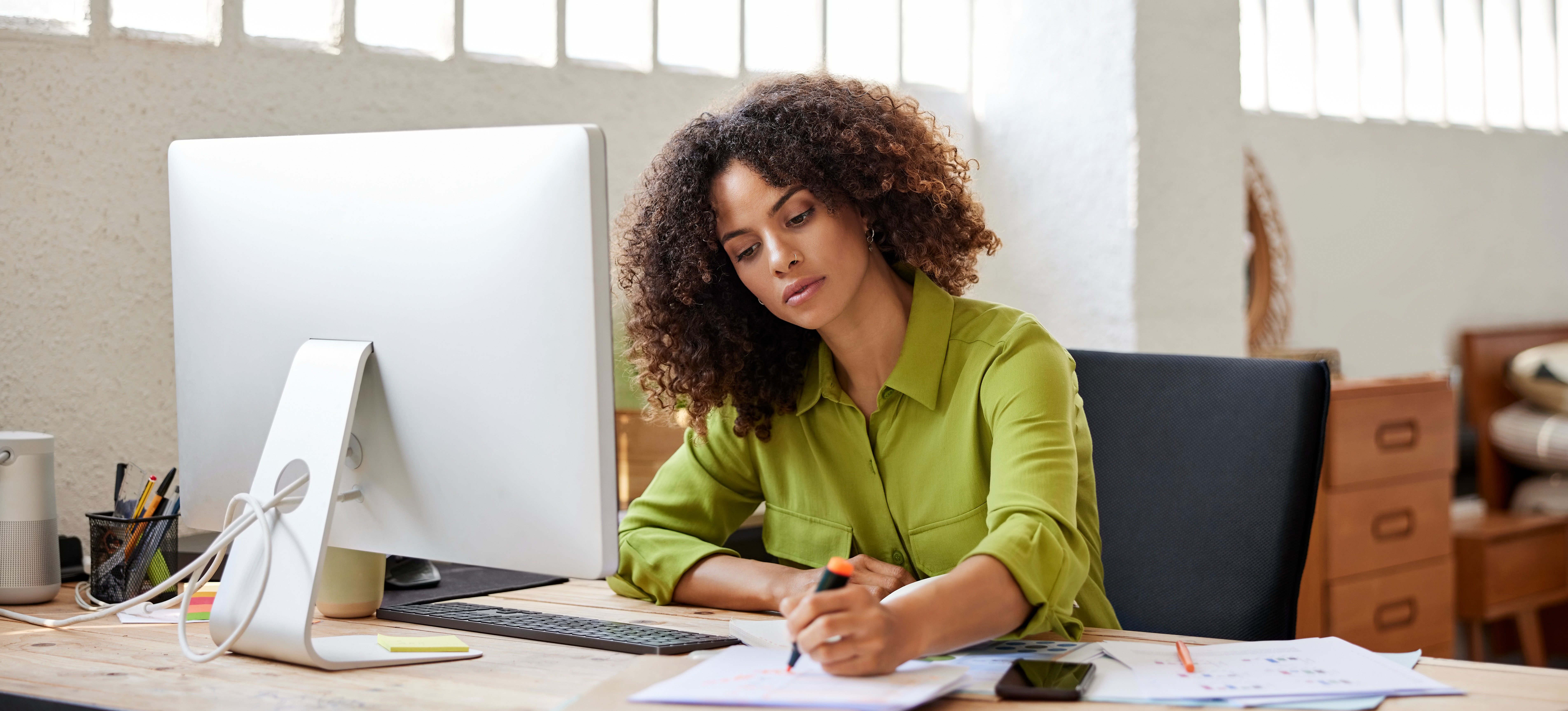 [Featured Image] A content creator in a sunny home office space holds a pen over paper as she sits in front of her computer monitor.
