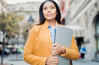 [Featured] A Doctor of Education student in a yellow jacket carries her laptop across a university campus.