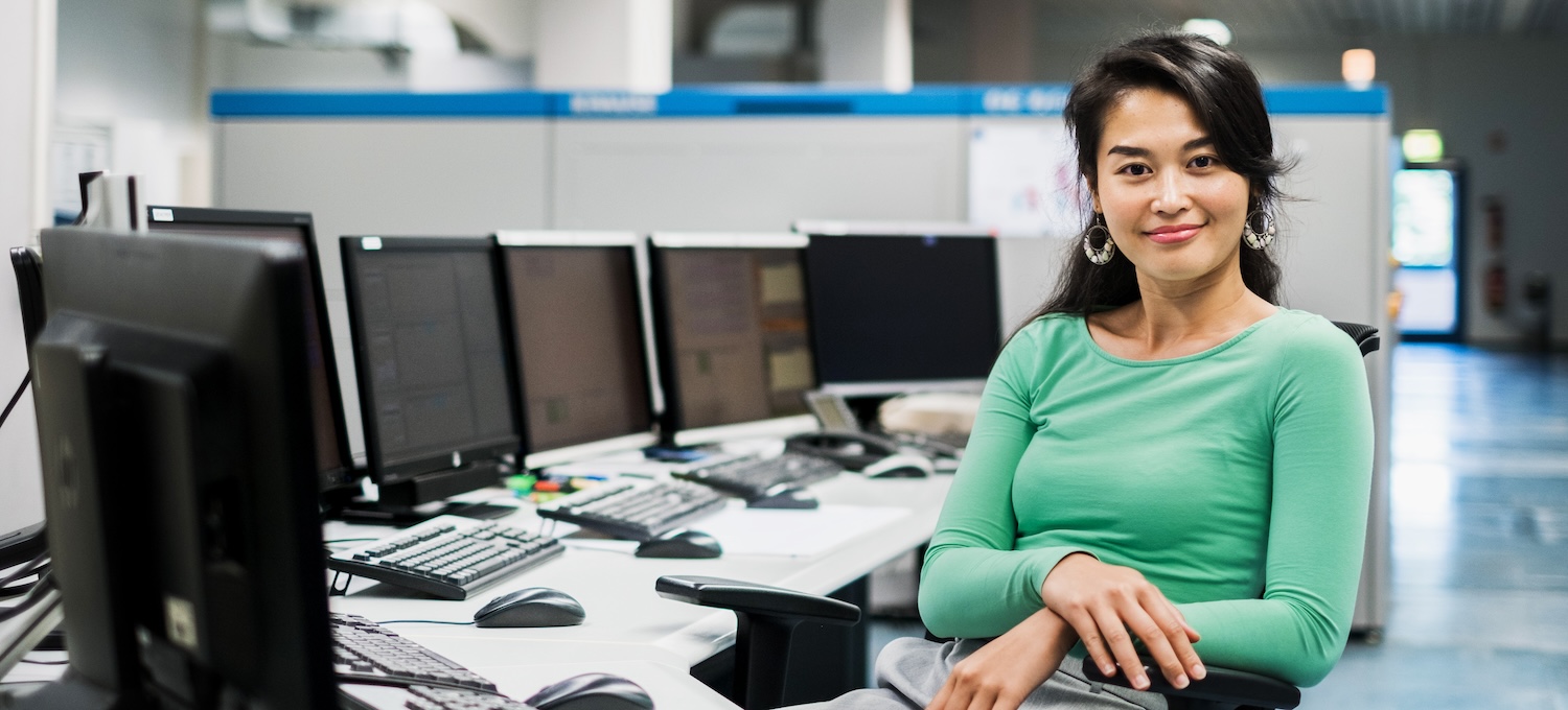 [Featured image] A woman in a green top sits in front of a bank of computers, smiling at the camera. 