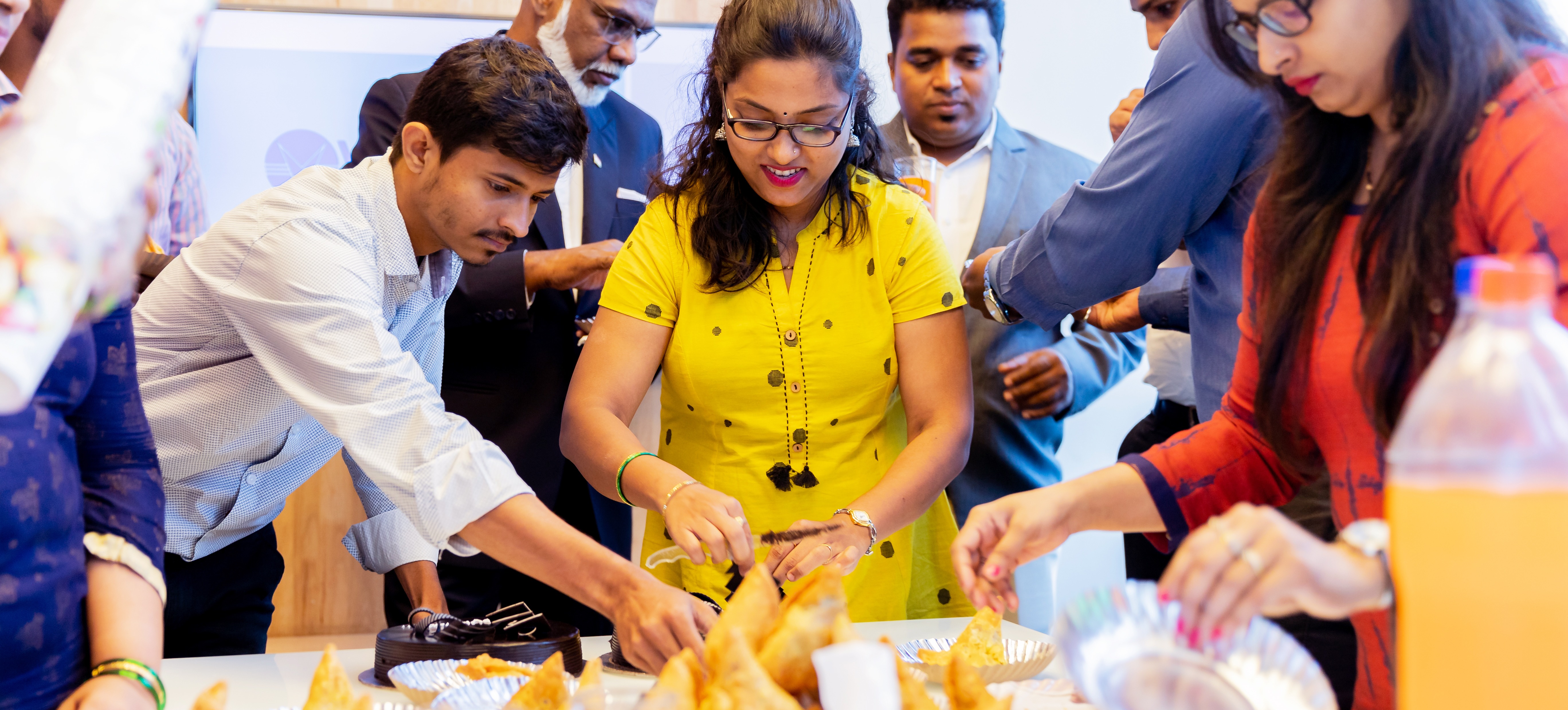 [Featured Image] An event manager displays how to do her hospitality job well by arranging food for a crowd of people who have assembled around a table and throughout the room.