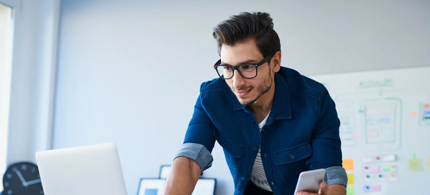 [Featured Image]: A machine learning enthusiast, wearing a blue jacket and glasses, stands in front of their computer, holding their phone.  