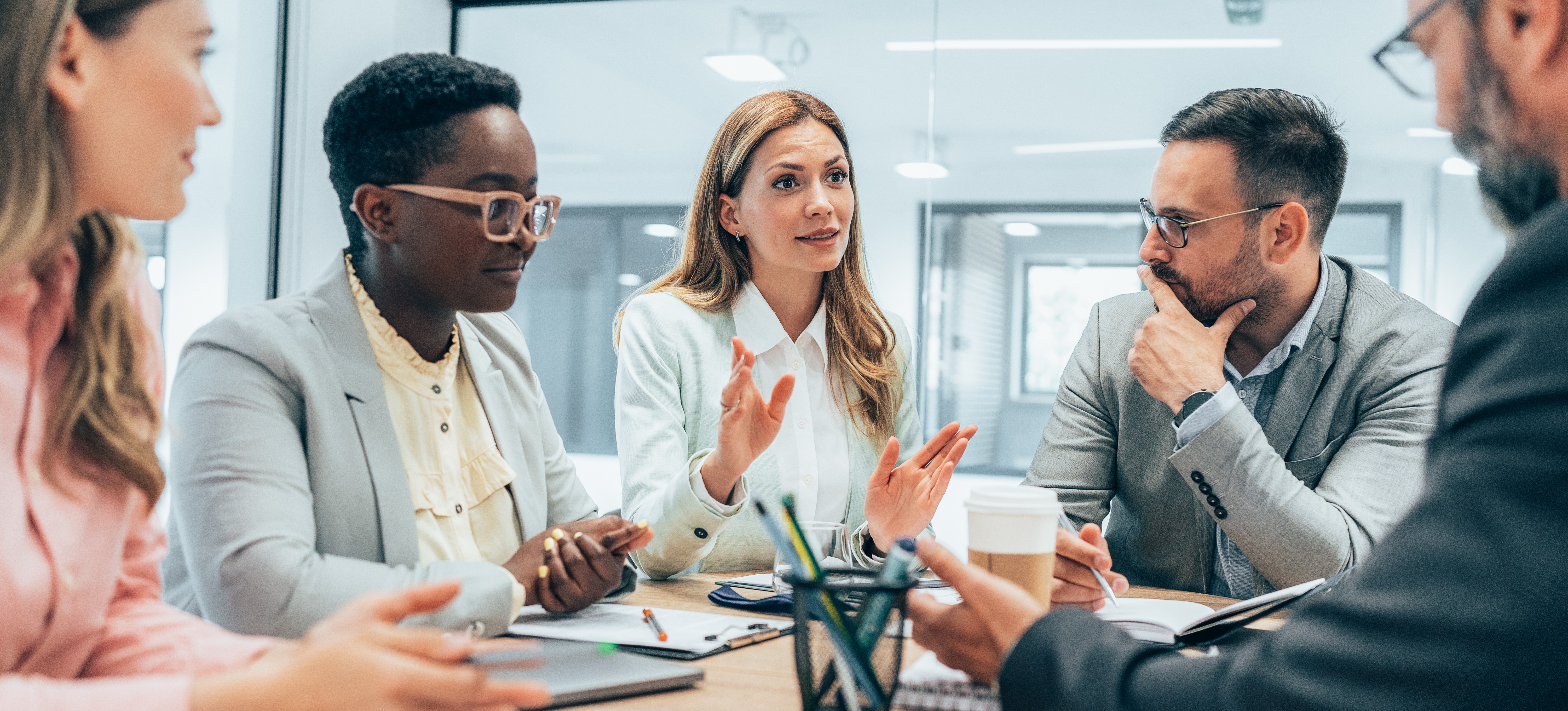 [Featured Image] A business person uses their people management skills to talk to four other team members in a business meeting.
