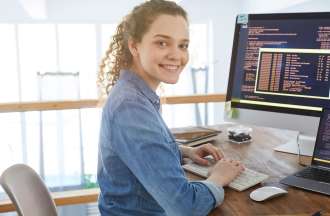 [Featured Image] A developer in a blue, collared shirt works on some basic computer programming at a desk with a laptop and desktop monitor.