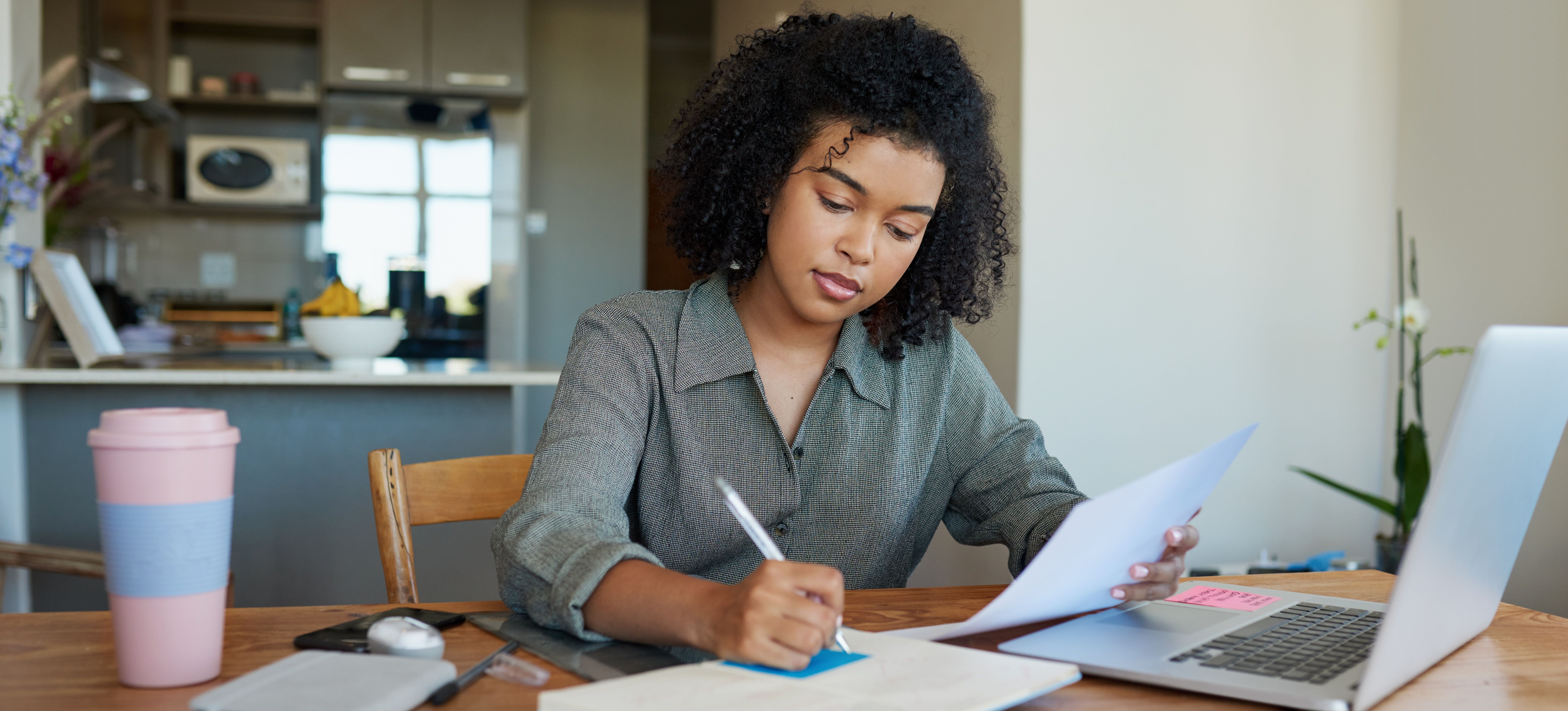 [Featured Image] A woman is writing on a sticky note and using her laptop.