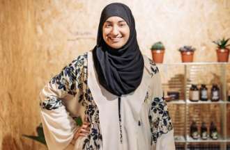 [Featured Image] A woman in hijab smiles and stands before the products she sells through her home business. 