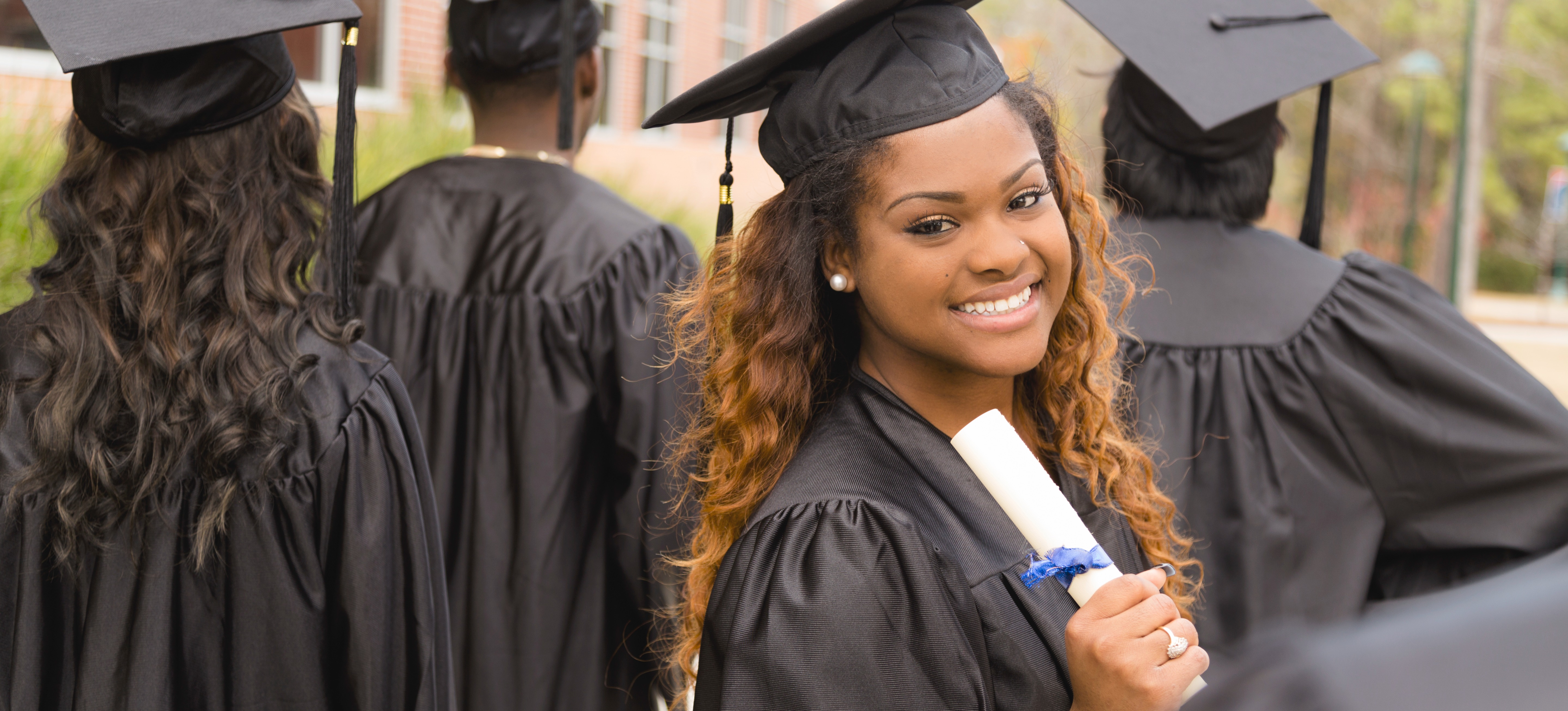 [Featured image] A graduate in their cap and gown holds their diploma and smiles after getting their bachelor’s degree with their classmates.