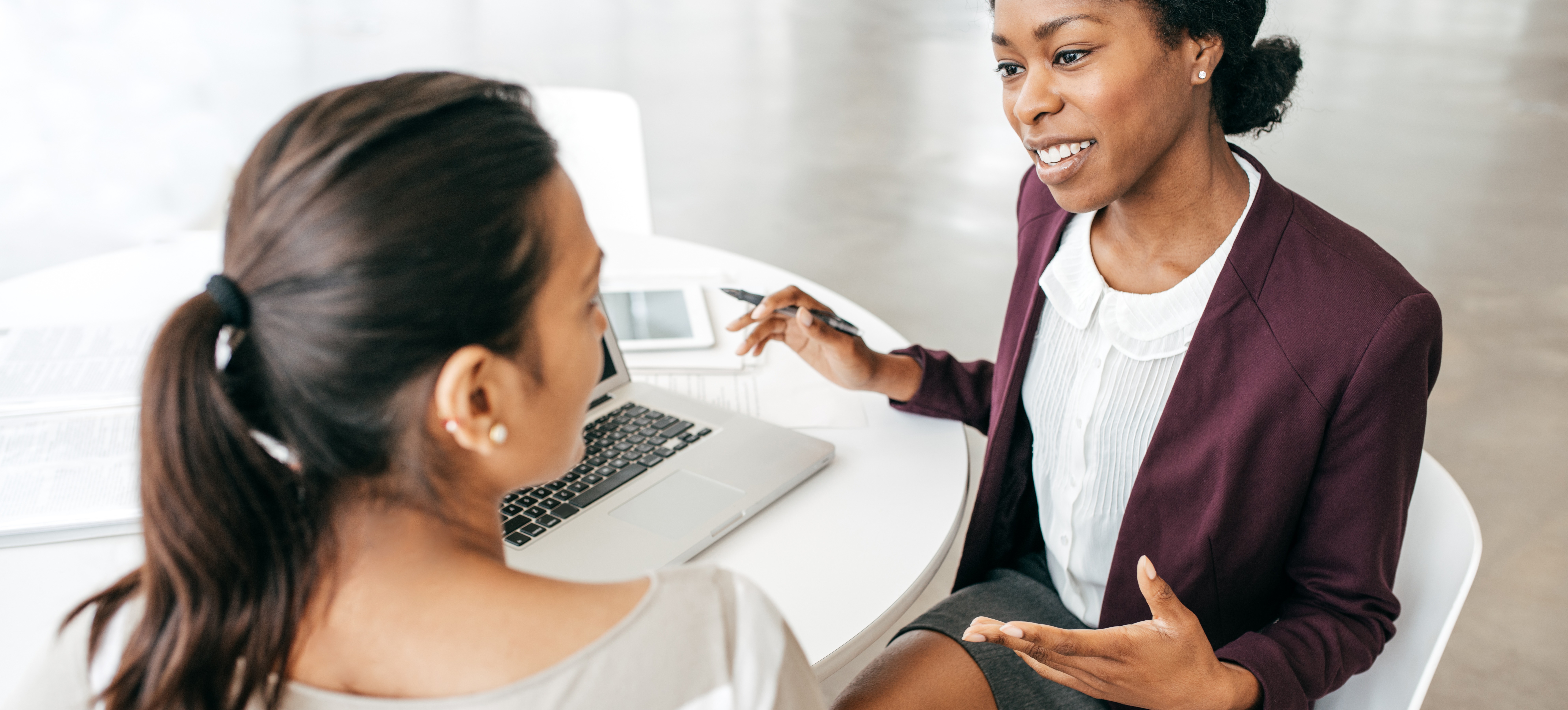 [Hauptbild] Während eines Vorstellungsgesprächs zwischen zwei Frauen, die an einem weißen Tisch sitzen, stellt eine Frau Fragen für ein Produktanalysten-Interview, um herauszufinden, ob ein Kandidat für die Stelle geeignet ist.