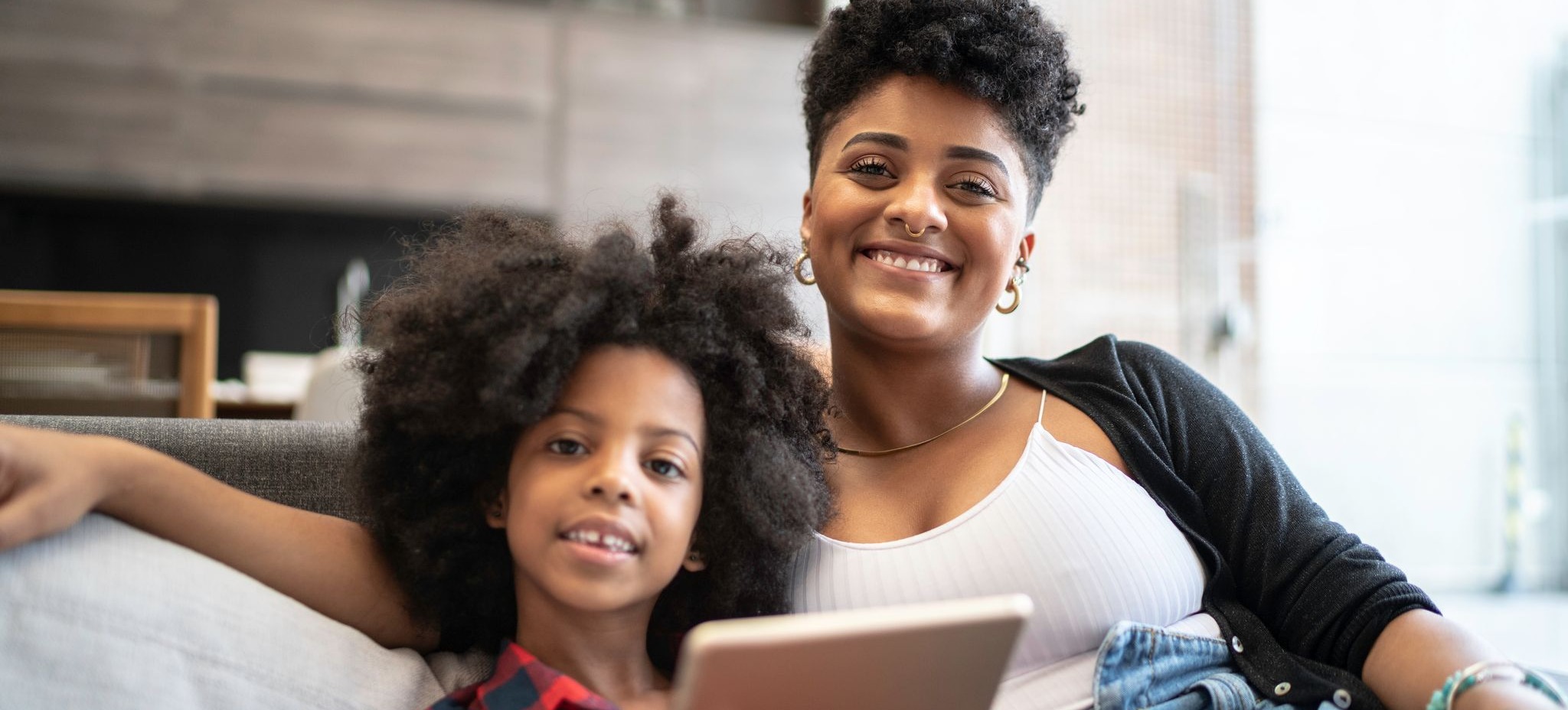 [Featured Image] A mother and daughter are at home sitting on a couch.  The daughter is holding a tablet.  