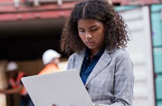 [Featured Image] A digital forensics examiner, one of many important cybersecurity in the maritime industry jobs, looks at a laptop while standing at a shipping port.
