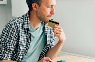 [Featured Image] A man in a plaid shirt is sitting in his house at a table, holding his phone and credit card and looking at his laptop, contemplating if should make the purchase in case it is a type of phishing.
