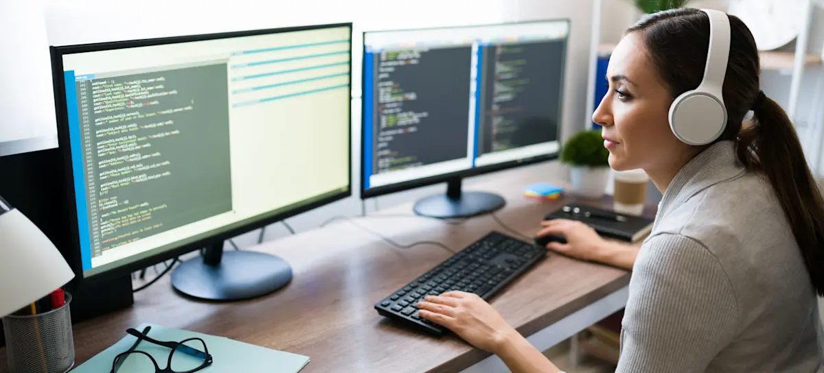 [Featured Image] A worker has headphones on and is looking at two computer screens that have Java programming language displayed.  