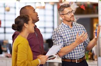 [Featured Image] Three coworkers discuss the contents of a whiteboard covered with post-it notes. 