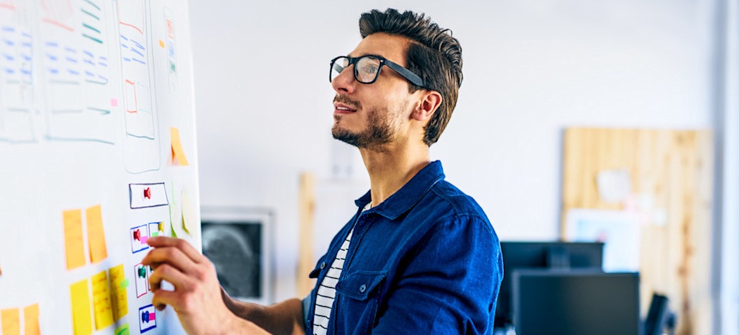 [Featured image] A UX designer wearing eyeglasses and a blue shirt draws a wireframe on a whiteboard.