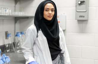 [Featured image] A PharmD student stands in a lab wearing a black headscarf, white lab coat, and blue medical gloves.