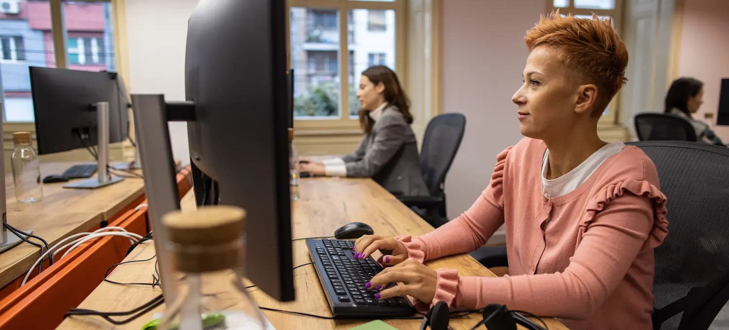 [Featured Image] A person at a computer desk in an office that is using peer-to-peer networking for all other computers and printers.