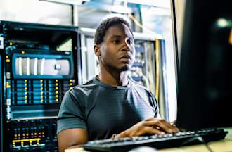 [Featured image] A CompTIA A+-certified IT worker sits in a server room and works on a black desktop computer.