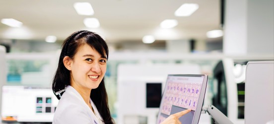[Featured Image]:  A female,  with long black hair, wearing a white uniform, is working in a lab, and is pointing to a computer screen, as she reads the charts and test results. 