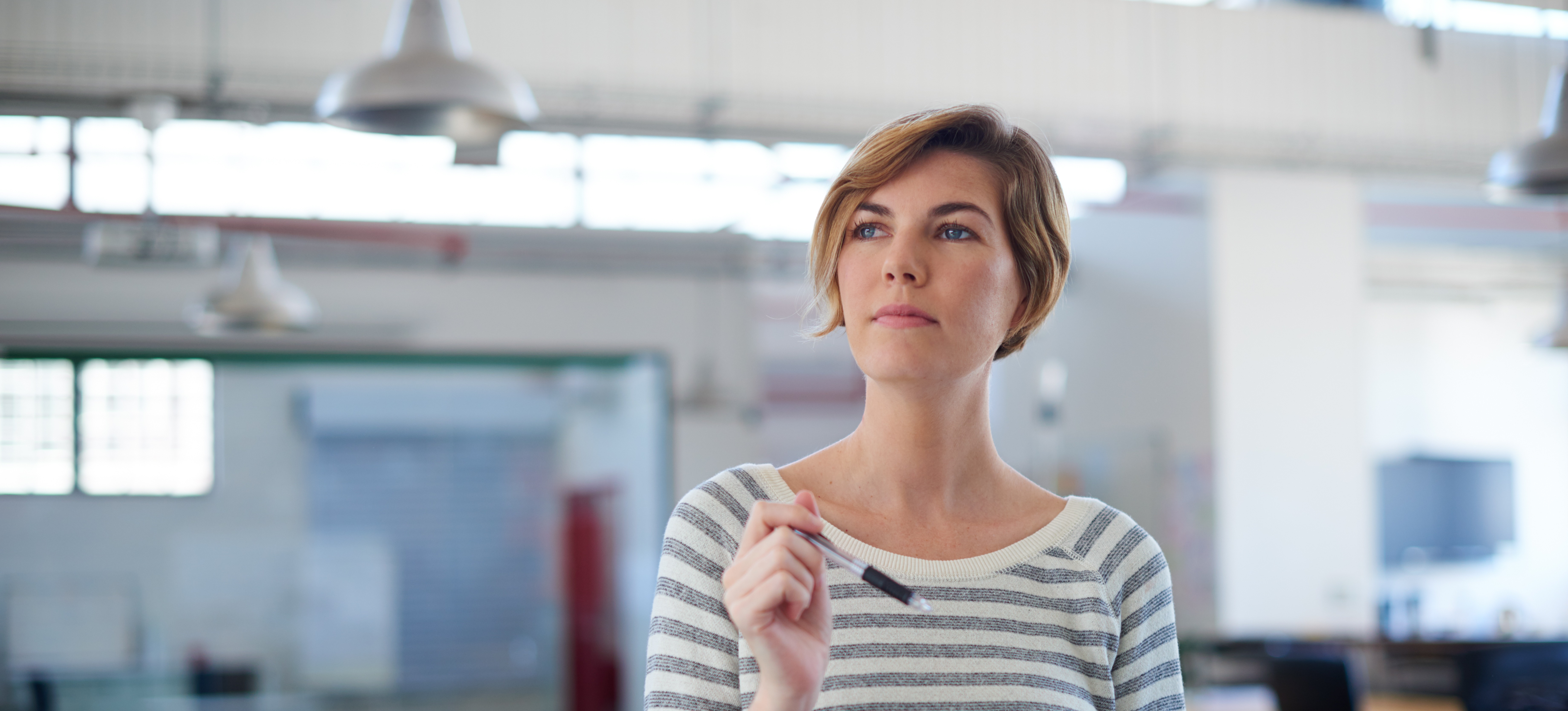[Featured Image]:  A product analyst with short blonde hair and wearing a white and gray striped top is holding a pen in one hand and documents in the other hand. She is standing near her desk in her office. 