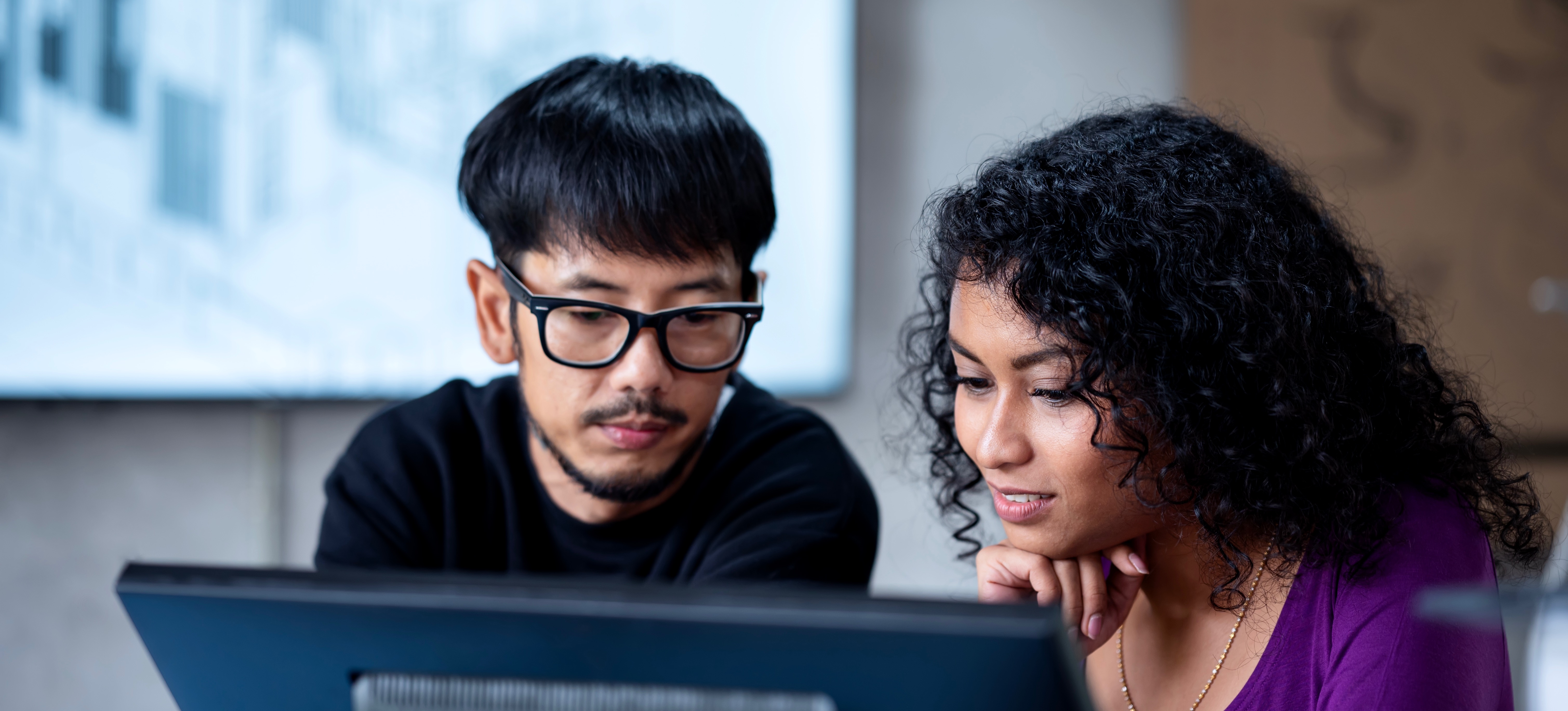 [Featured Image] A woman working in software project management sitting behind a computer, explaining the project's scope to a coworker.