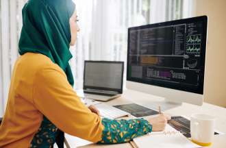 Female data engineer sits in front of a dual computer screen looking at data visualizations and writing in a notebook