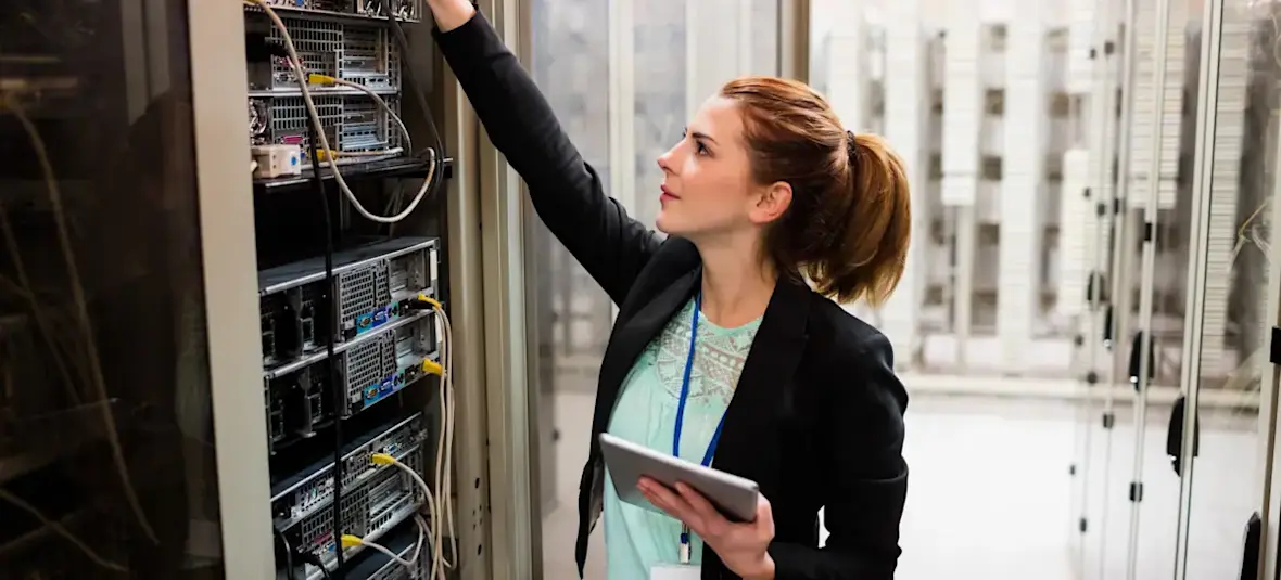[Featured Image] In a server room, a network operations engineer examines a server while holding a tablet.
