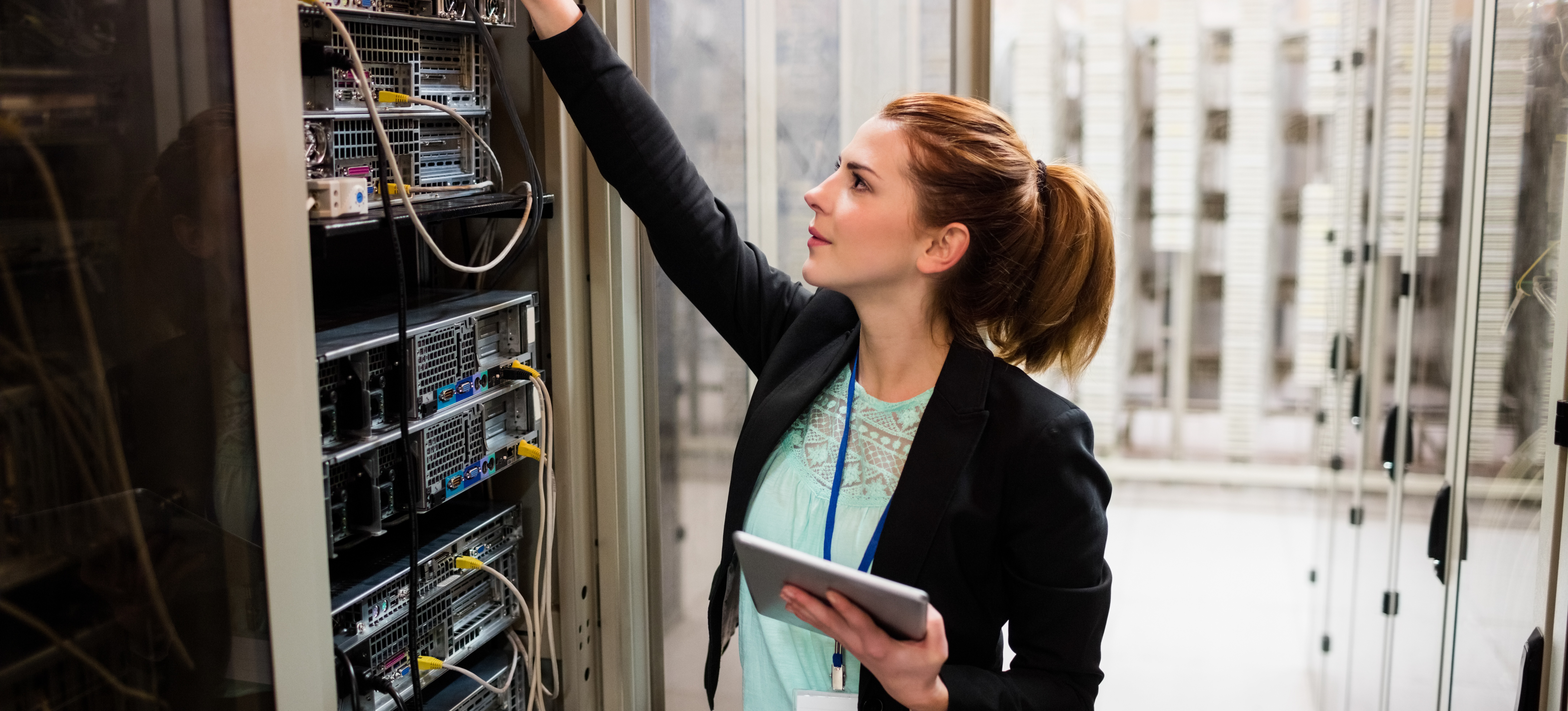 [Featured Image] In a server room, a network operations engineer examines a server while holding a tablet.
