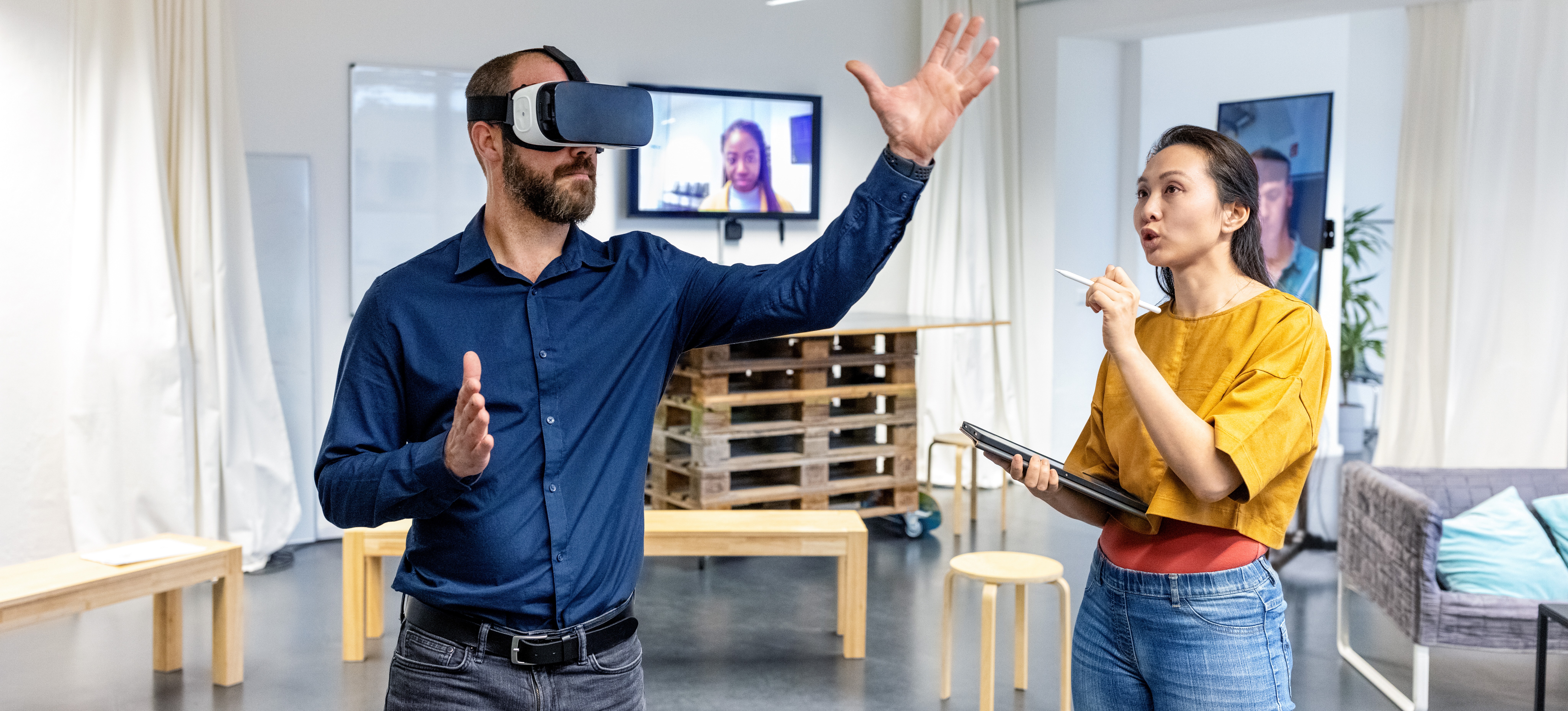 [Featured Image] A businessperson teaches the difference between extended reality versus virtual reality while their co-worker wears a VR headset beside them.
