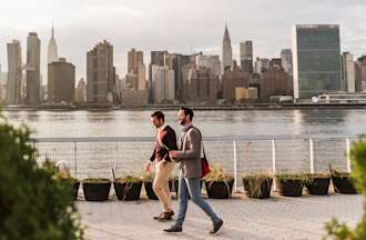 [Featured image] Two mean in business attire walk along the waterfront with the New York City skyline in the background.