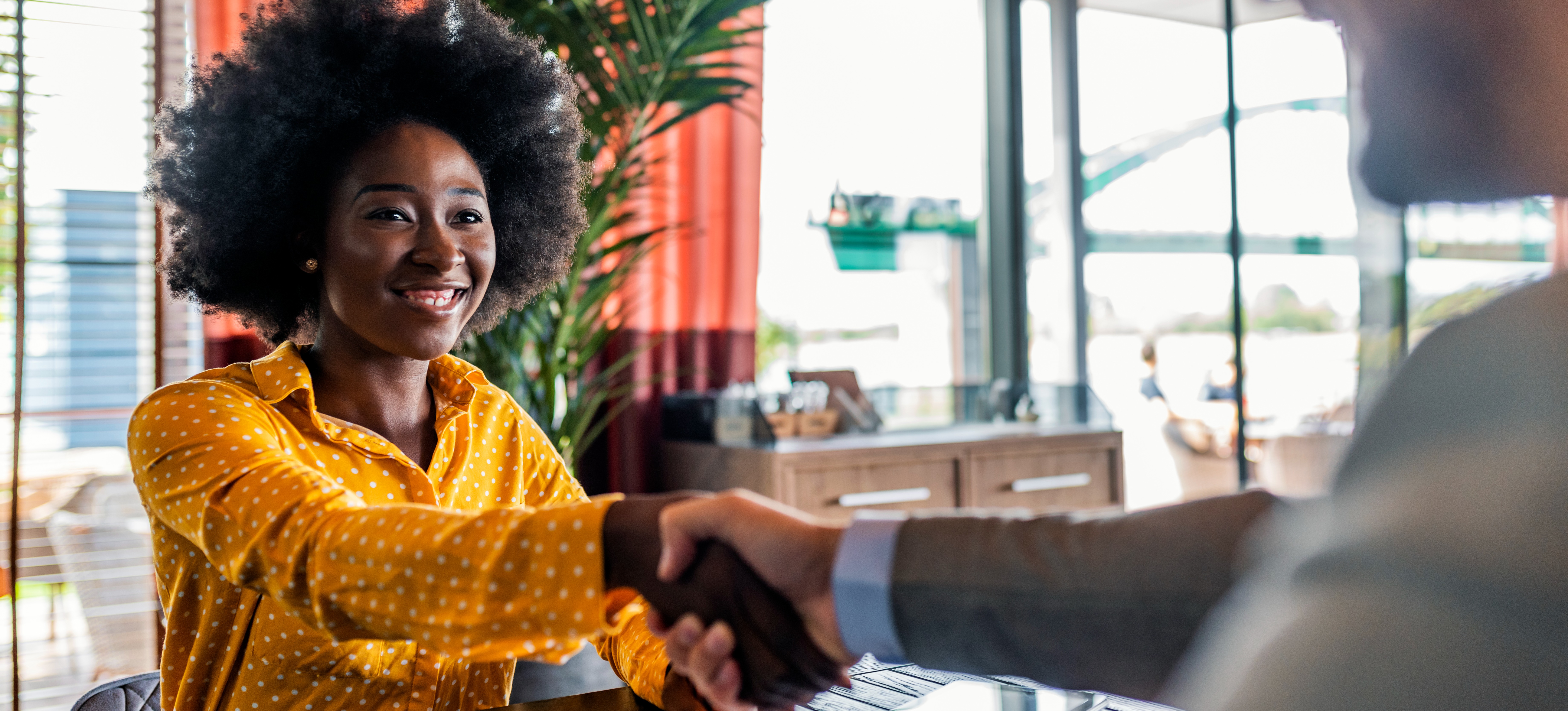 [Featured Image] A young woman who learned how to let recruiters know you're open on LinkedIn shakes hands with a job interviewer across a desk.
