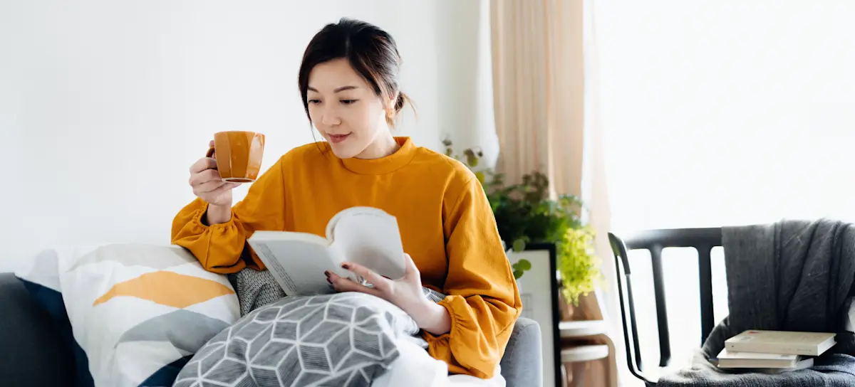 [Featured Image] A person is sitting on their couch, drinking coffee while reading a book, and practicing work-life balance.