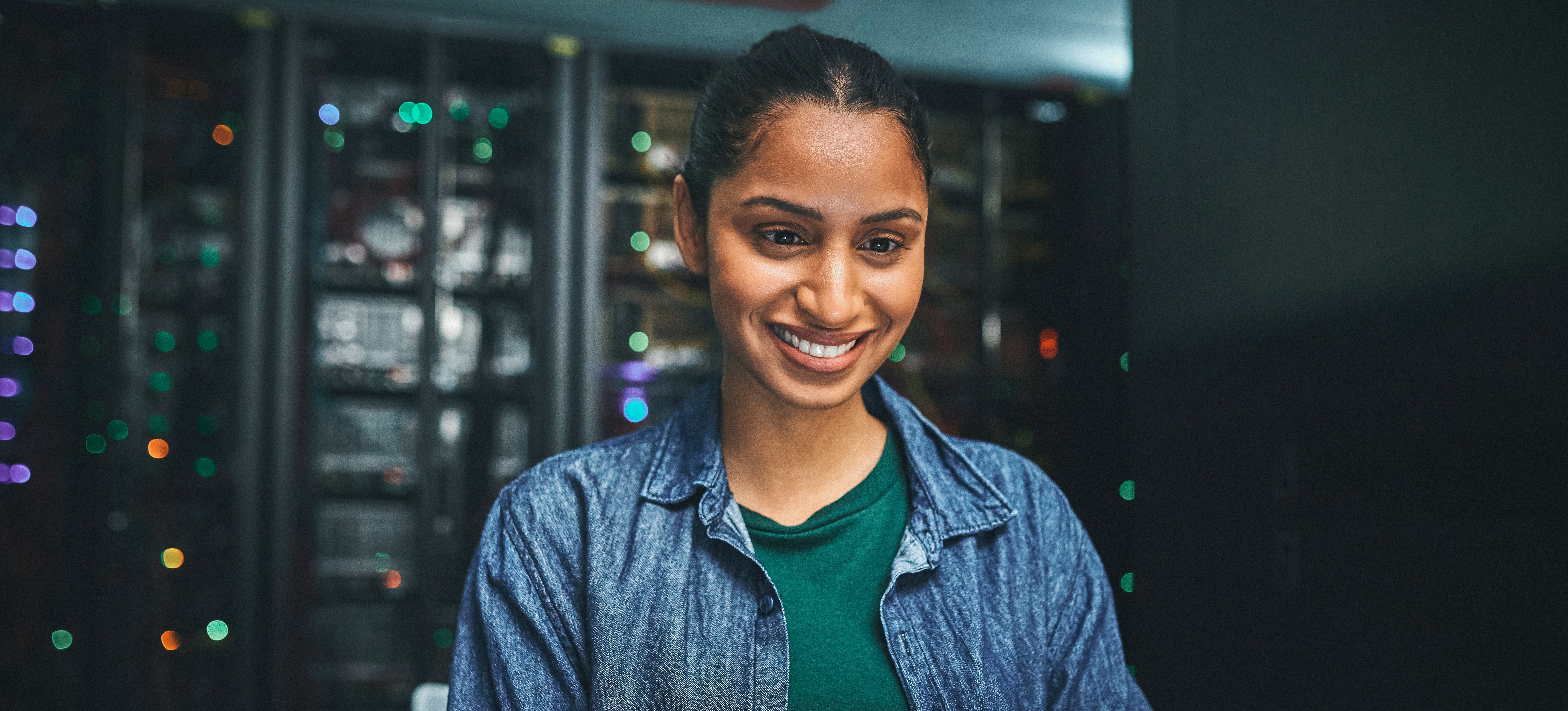 [Featured Image] A smiling security architect reviews an organisation’s cybersecurity measures on a laptop while sitting in a server room.
