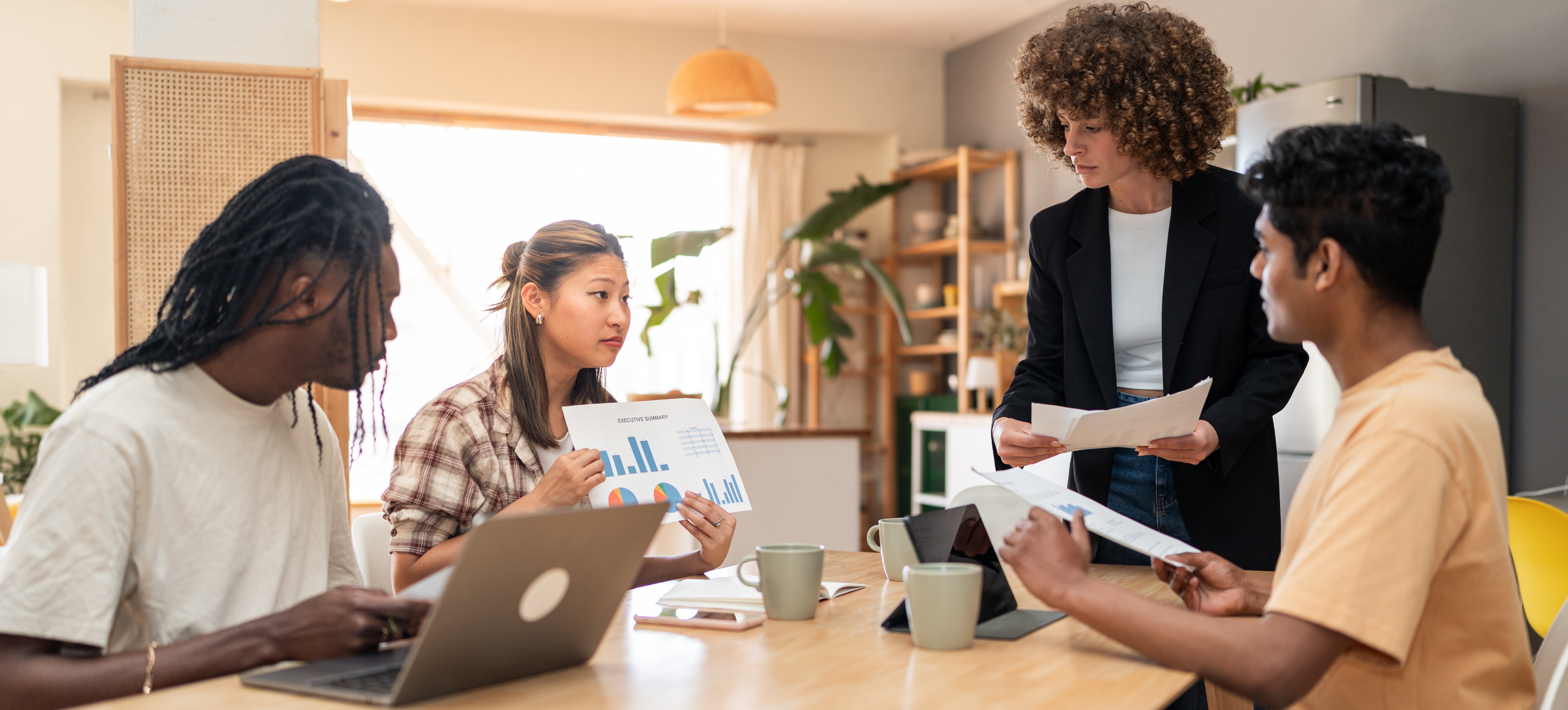 [Featured Image] A group of four colleagues is gathered around a wooden table in a brightly lit contemporary office space. They are viewing a chart that is being presented by one of the group members. A laptop and notebooks are visible on the table.
