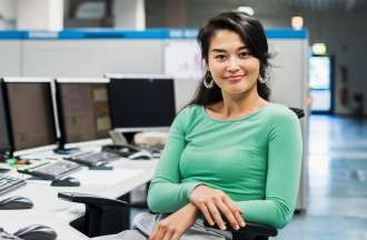 [Featured image] A young woman wearing a green long-sleeved top sits in front of several desktop computers. She smiles at the camera. 