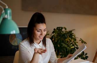 [Featured Image] An executive assistant is holding papers and working at her desk with her laptop. 
