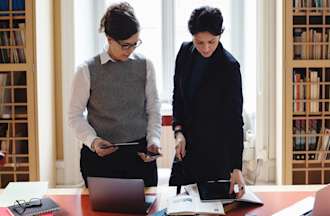 [Feature Image] Two people stand in front of a conference table and review accounting numbers in a library.