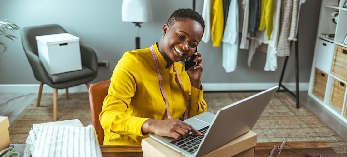 [Featured Image] A smiling small business owner sits in their tailor shop and creates a Google shopping campaign on their laptop.