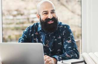 [Featured Image] A person with a beard sits at a desk in front of a laptop smiling. 