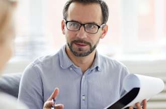 [Featured Image] A project manager in a blue button-down shirt holds a clipboard and pen and interviews a candidate. 