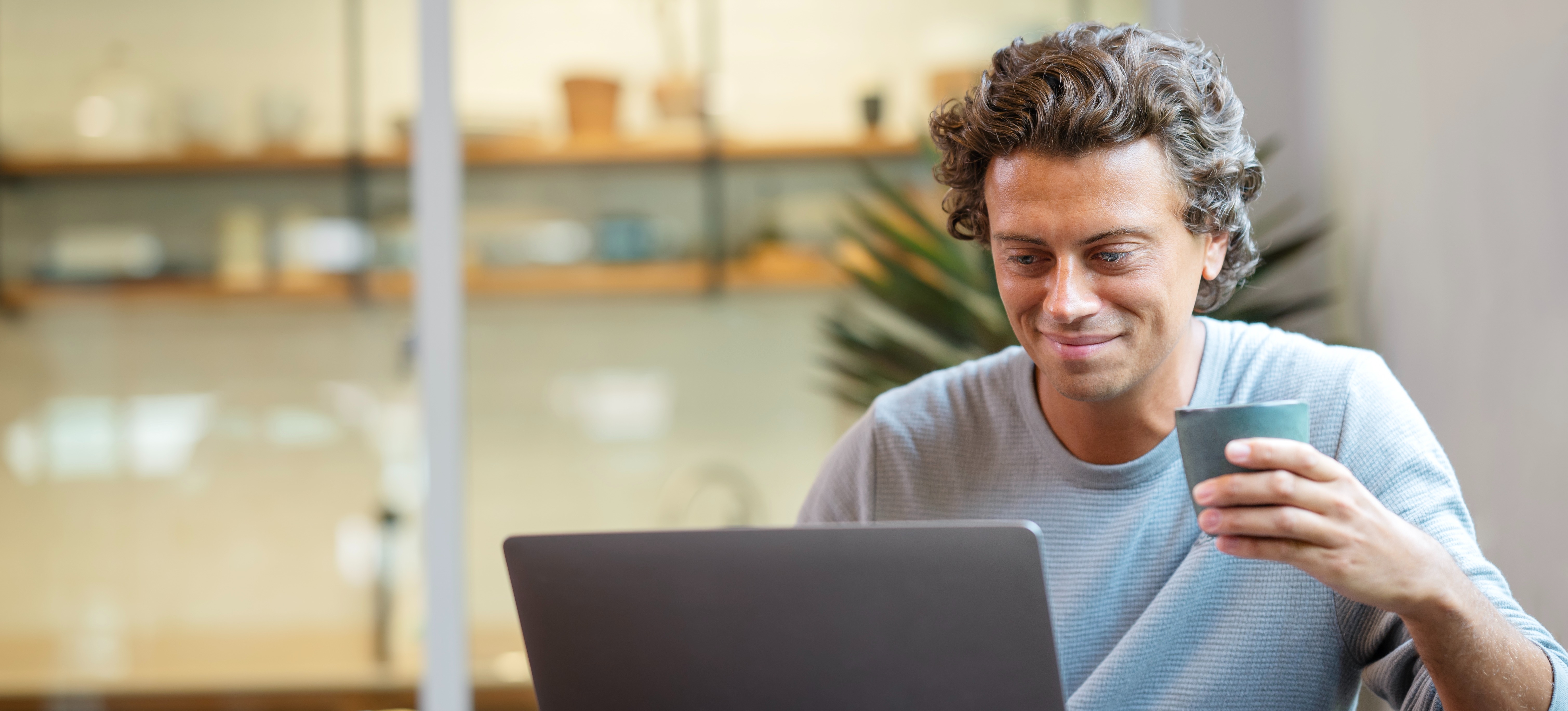 [Featured Image] A smiling businessman in casual clothes and holding a cup of coffee works from his living room, confident that his organization's cloud solutions will enable him to work remotely with ease.
