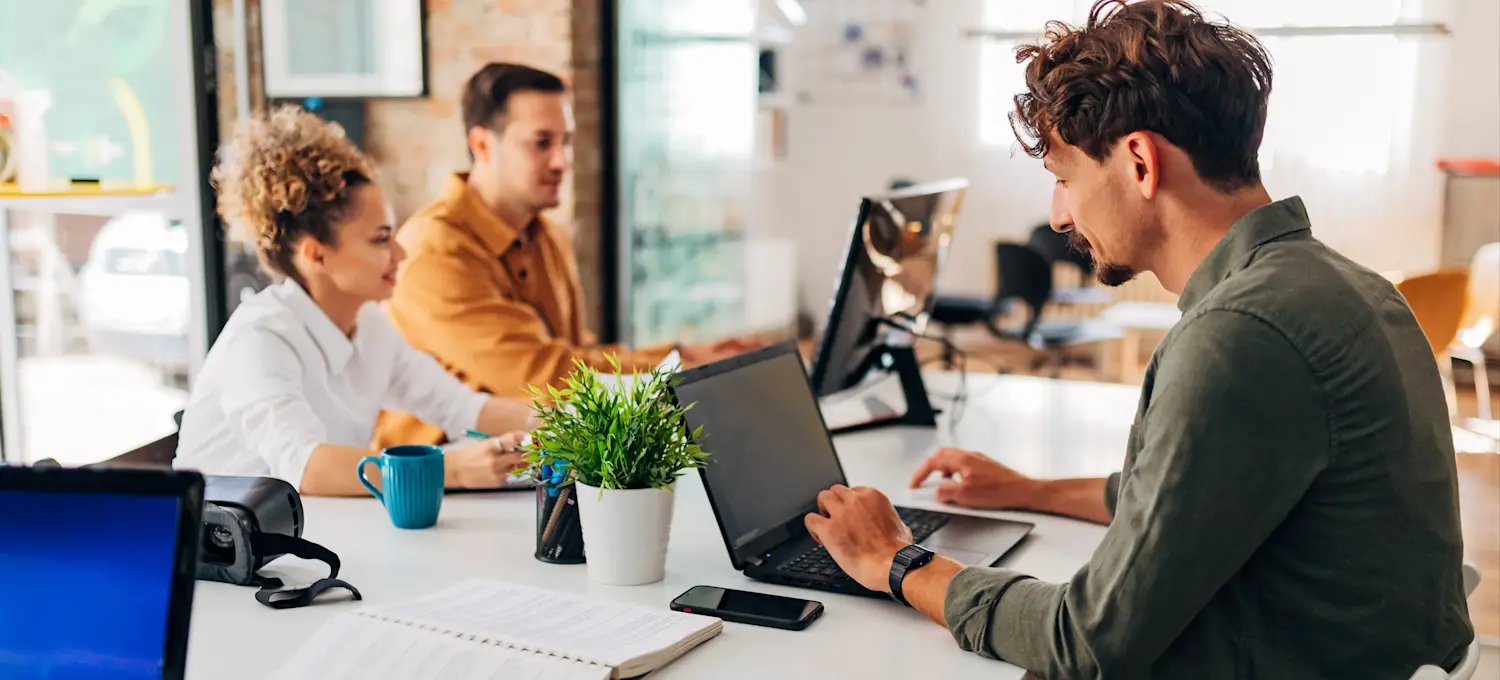 [Featured Image] Three web developers sitting at a table in an open concept office use backend languages to write code on their laptops.
