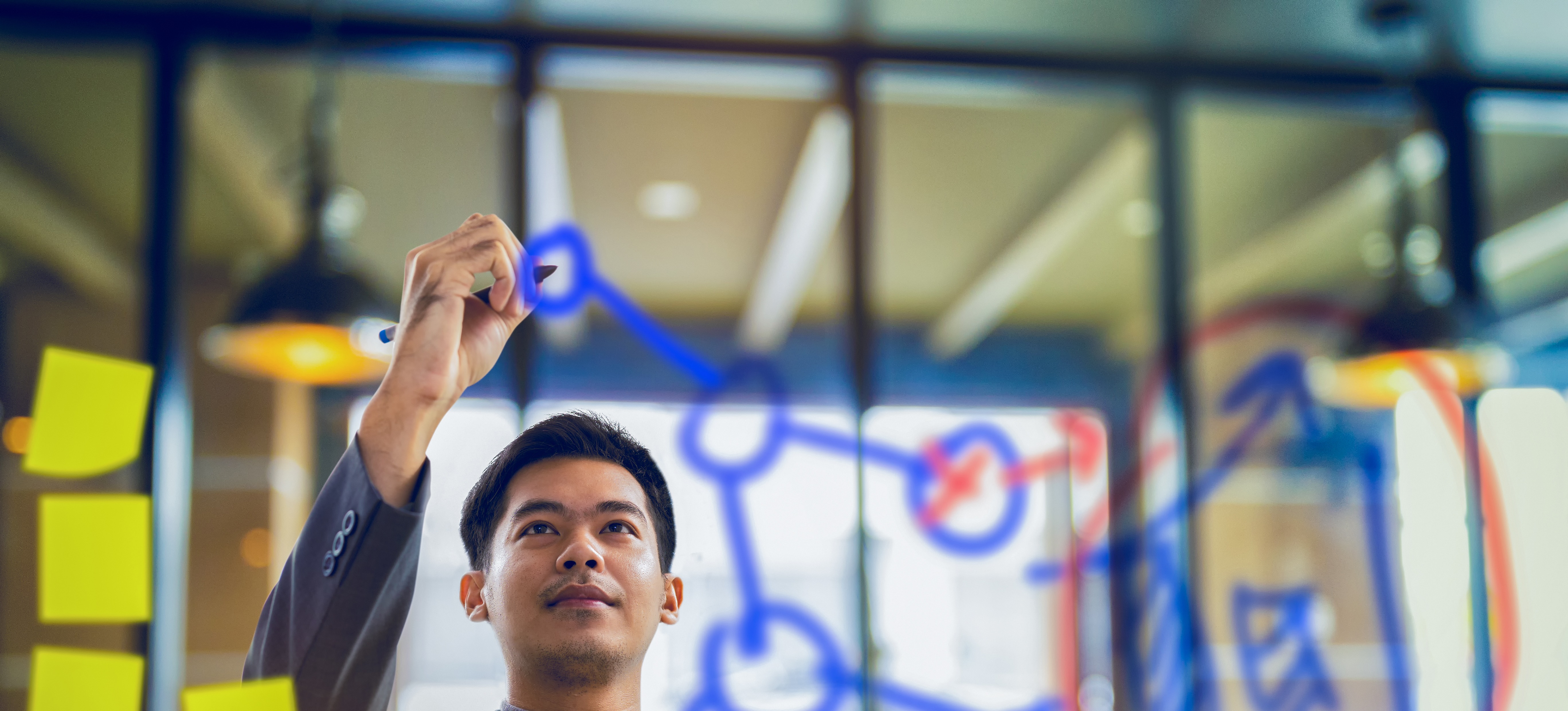 [Featured Image] A supply chain analyst draws a diagram on a window in blue ink. There are yellow Post-it notes stuck to the left side of the window. 