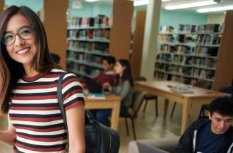 A smiling business school student in a striped dress stands in a library with her phone in her hand.