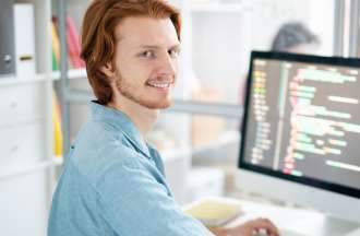 [Featured image] A person with short red hair and a beard, sitting in front of a desktop computer with code, smiles. 