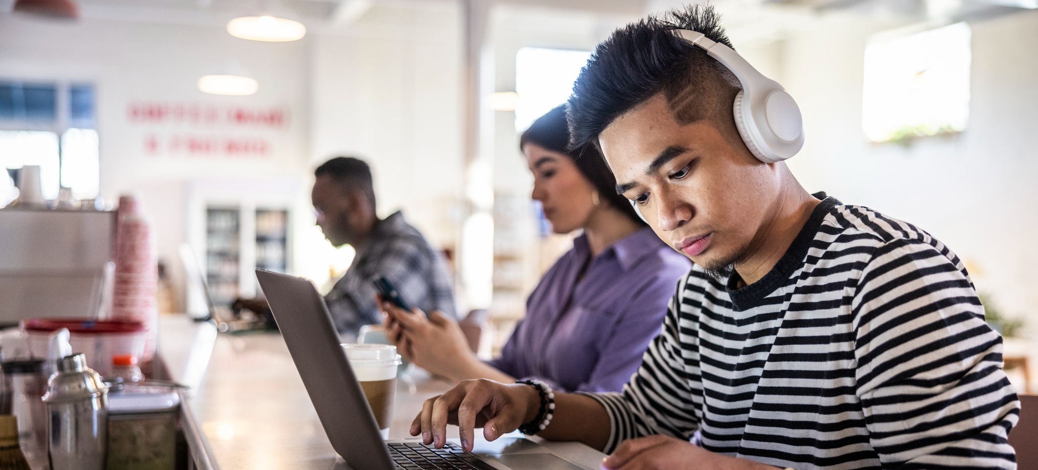 [Featured Image] A learner in an online master’s in computer science program studies and completes assignments on their laptop in a coffee shop while wearing headphones.
