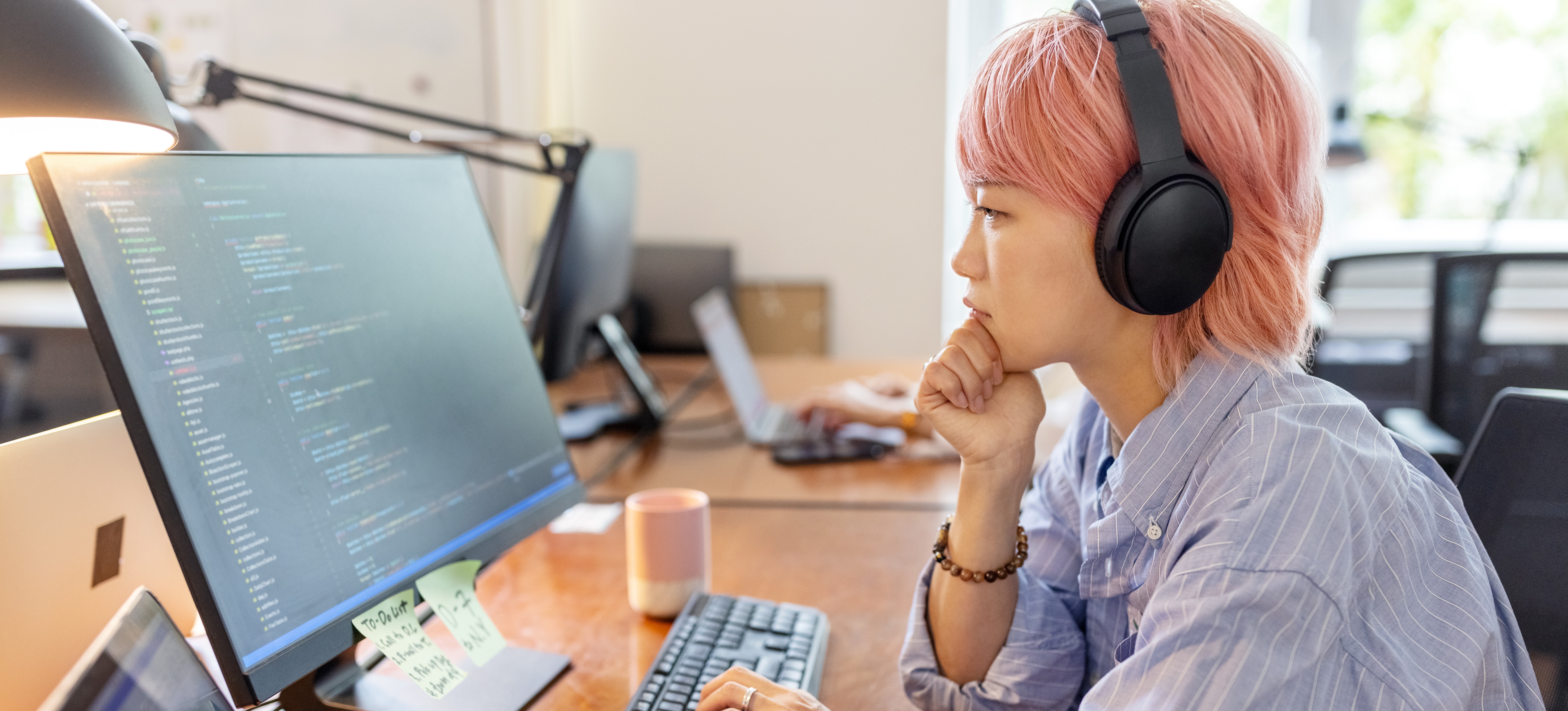 [Featured image] Side view of a data analyst working at their computer. They are looking at the computer monitor, which appears to have code on the screen, and thinking as they work in their office space. 
