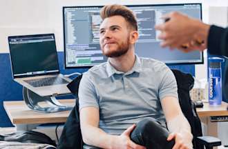 [Featured Image] A machine learning engineer in a gray shirt sits with his back to his desk and speaks with co-workers.