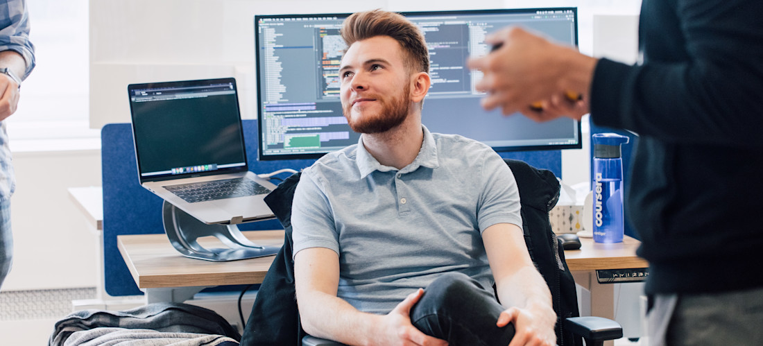 [Featured Image] A machine learning engineer in a gray shirt sits with their back to their desk and speaks with co-workers.
