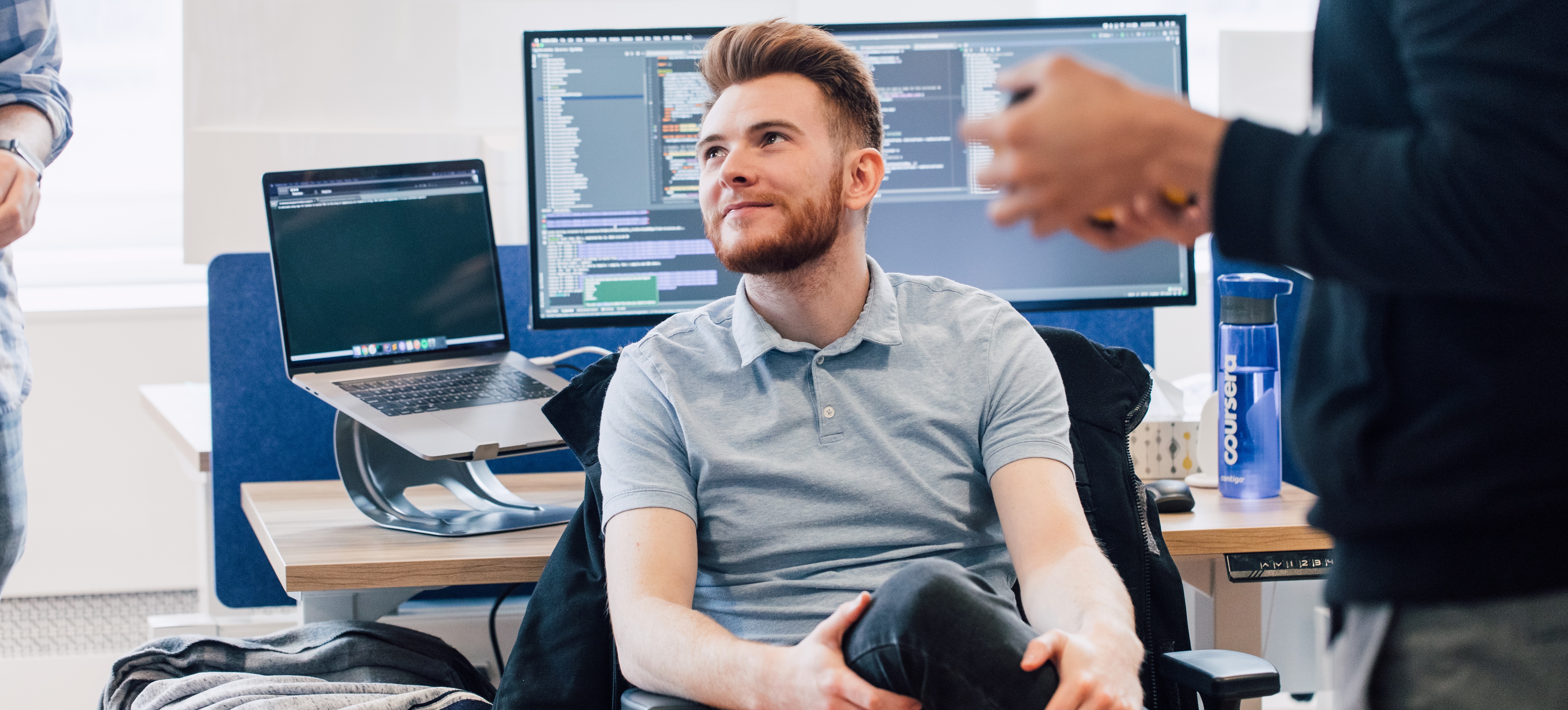[Featured Image] A machine learning engineer in a gray shirt sits with their back to their desk and speaks with co-workers.